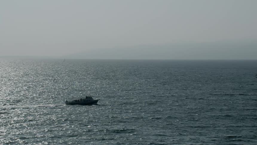 A boat with several people onboard sails across the water among sailboats as the sunlight reflects the ripples in the waves