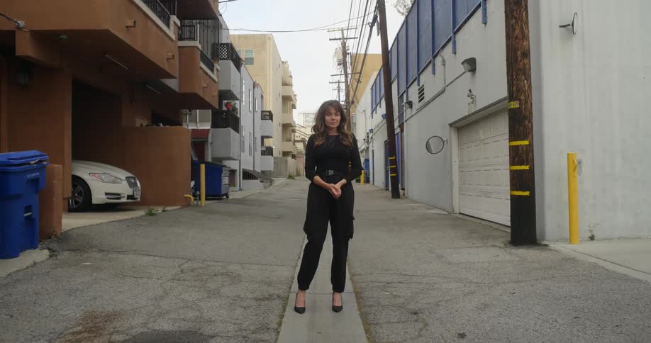 Woman with brown hair black shirt pants and high hills stands in the narrow street