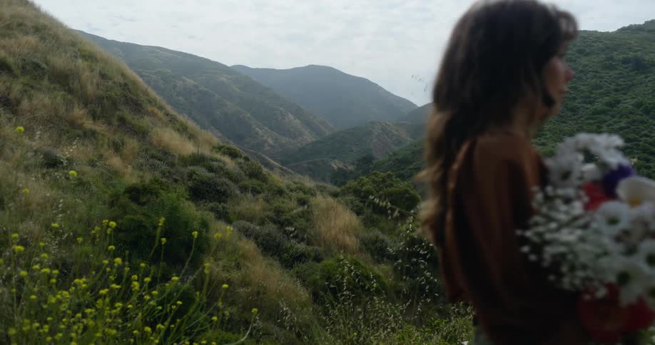 Woman with brown hair and blouse carries a bunch of flowers in mountainous area