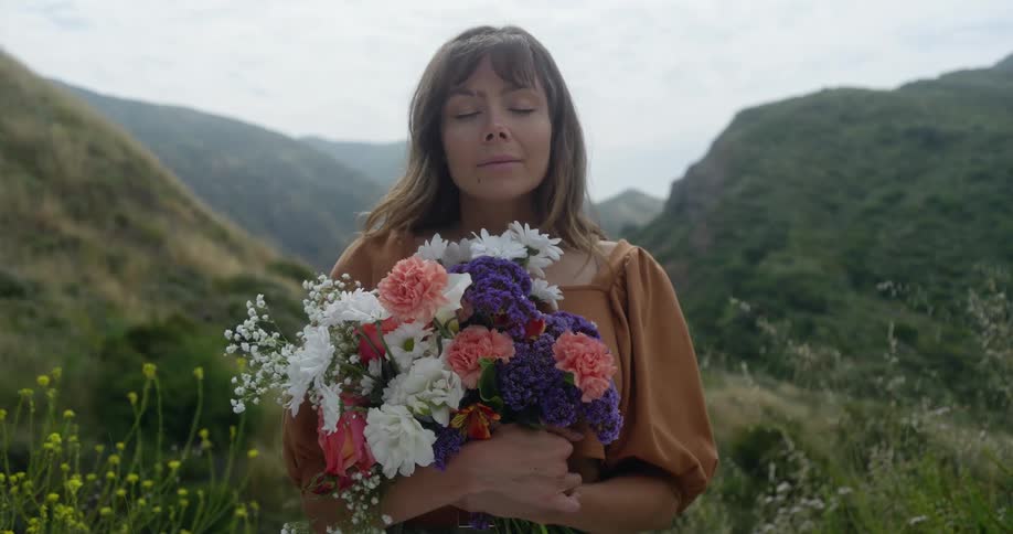 A woman standing outdoors holding a bouquet of flowers with mountains in the background behind her