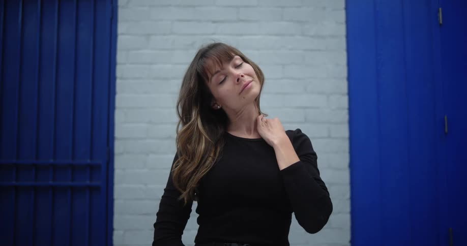 A woman is standing in a doorway with a brick wall behind her as she pushes back her long brown hair with her hand and places her other hand on her wrist as she looks ahead