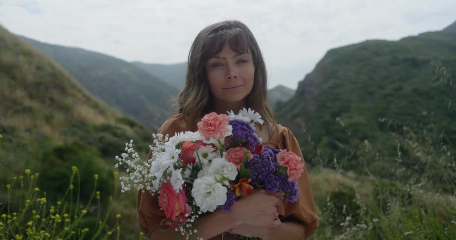 Woman holding a bunch of colorful flowers looks at camera in mountainous area