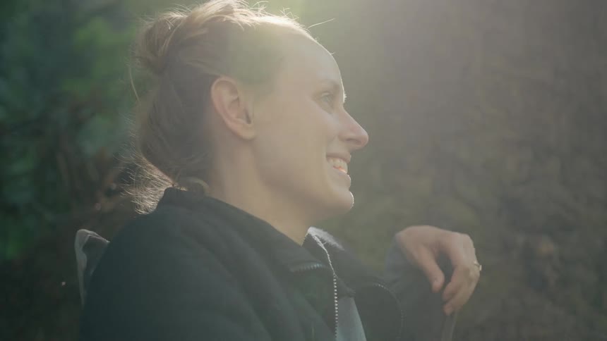 The sun shines brightly through the trees as a woman is standing in the woods smiling and laughing