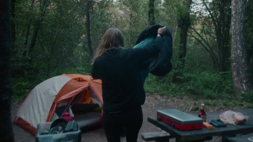 A woman is carrying a sleeping bag to a tent in the woods