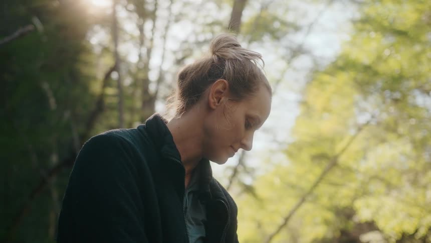 Blonde woman with black coat sits on tree stump in the forest smiling