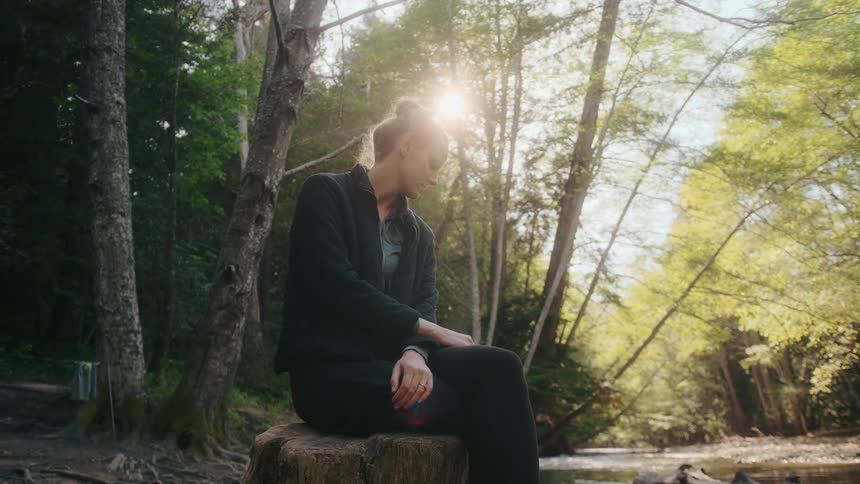 Blonde woman with black jacket and pants sits on tree stump in the forest