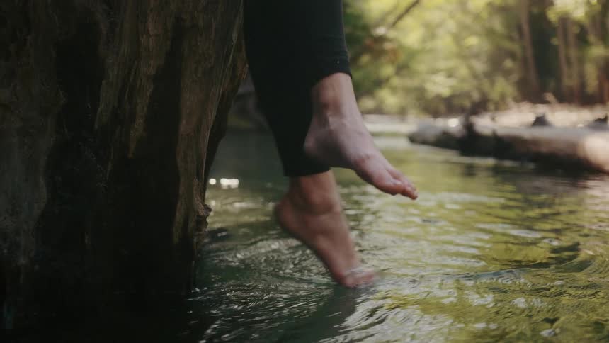 Two feet dangling slightly over a creek one of which dips their toes in the water