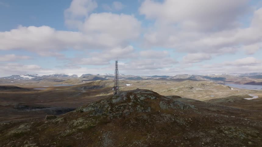 A utility tower stand on top of a mountain with more mountains in the distance under a cloudy sky