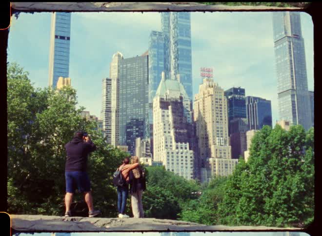 Two people standing on a rocky ledge holding each other looking at a city skyline as a person taking a picture walks up behind them