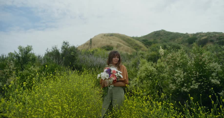 Woman standing among tall green vegetation while she holds a bouquet of colorful flowers
