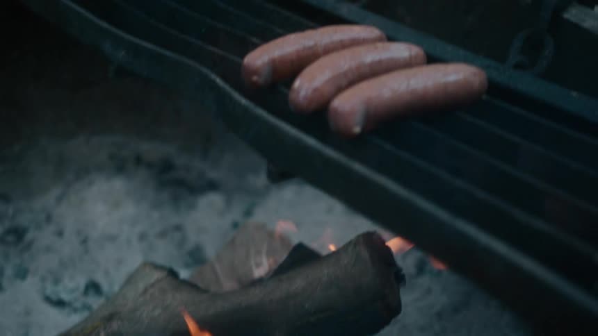 A pile of wood burns while grilling three sausages above it