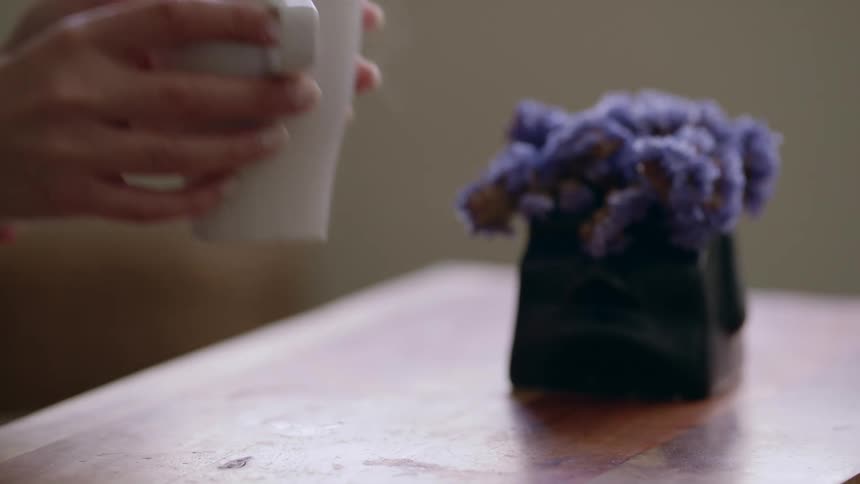 A close up of a woman grabbing a white coffee cup off a table near a purple flower