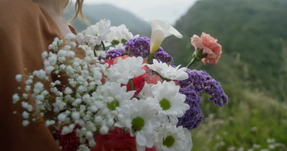 Woman stands in meadow near mountains on a misty day and holds a bouquet