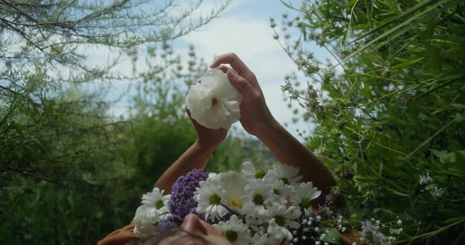 A woman is lying on the ground outside with a bouquet of flowers on her chest as she hold up a white flower with both hands