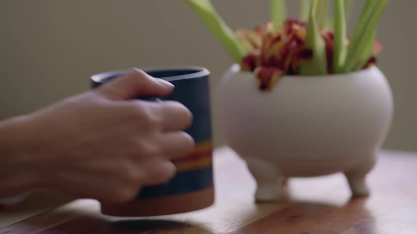 A close up of a mug on a table when a person grabs it and picks it up off the table