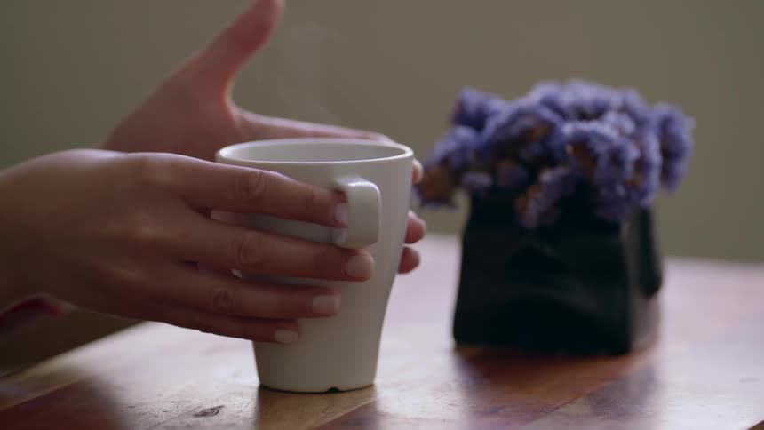 Close up of a table with purple flowers on it as a person places a white mug with a hot beverage in it on the table