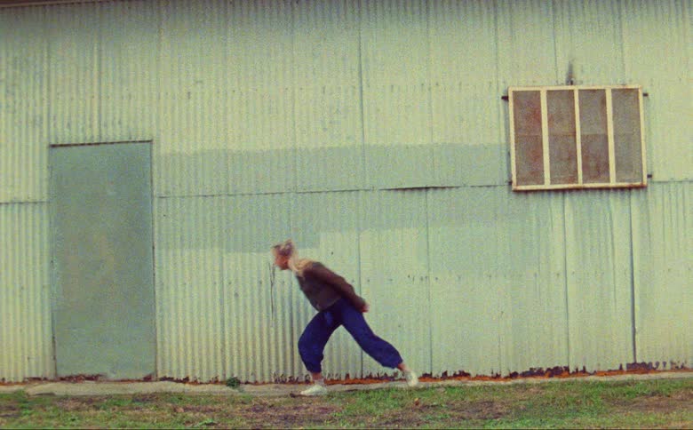 Blonde woman with transparent shirt blue pants dances in front of corrugated metal wall