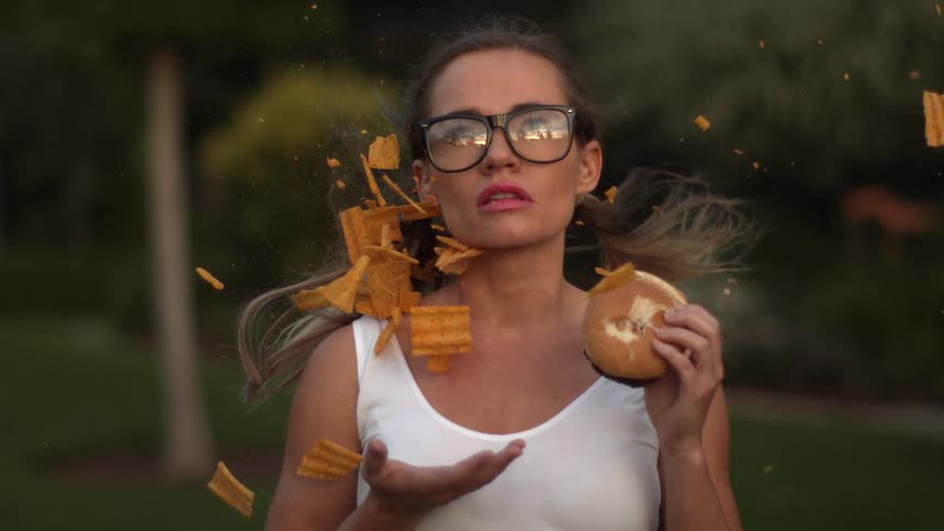 A woman holds a hamburger while a plate of chips is blown back into her face in extreme slow motion
