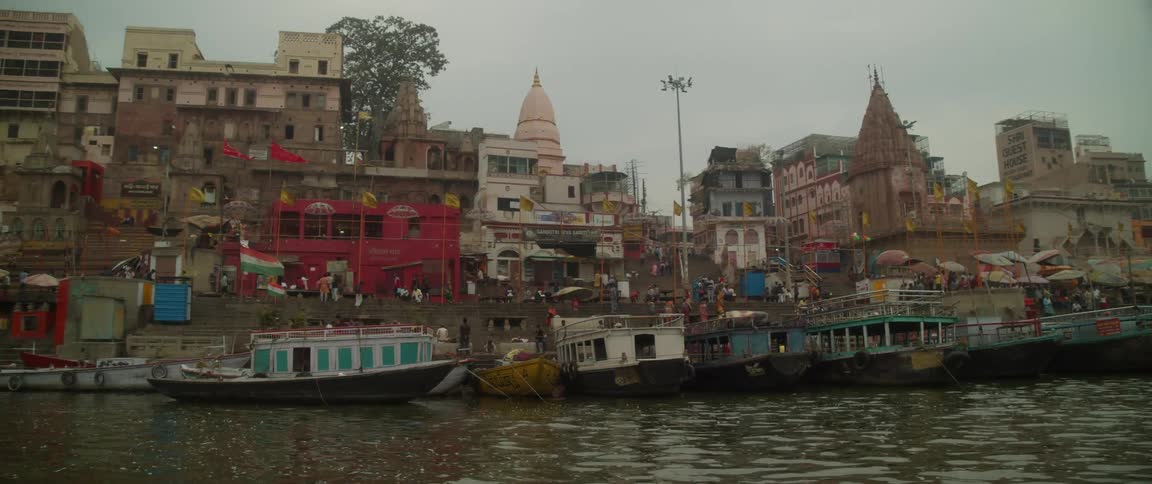 Boats are docked in the water along the coast as people walk on the streets among buildings and temples with beautiful architecture