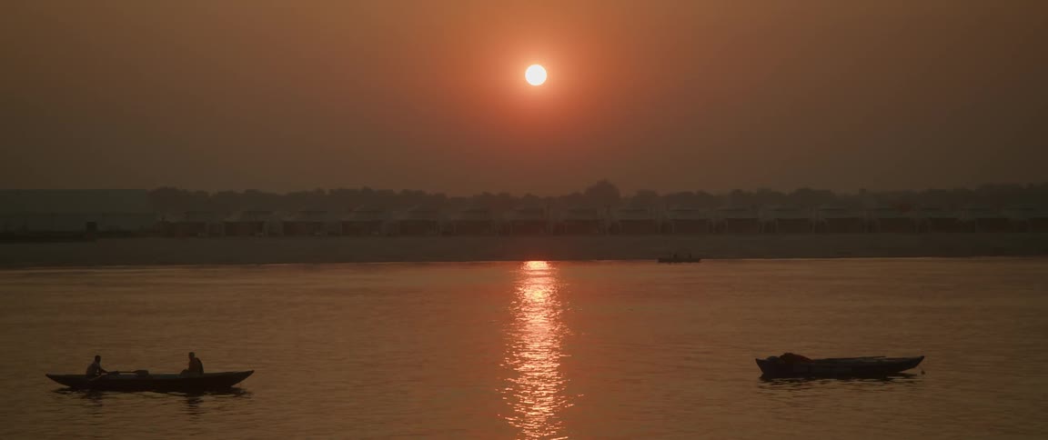 Two people sail in a small rowboat and another small boat floats on the water as the sun sets in a deep orange sky