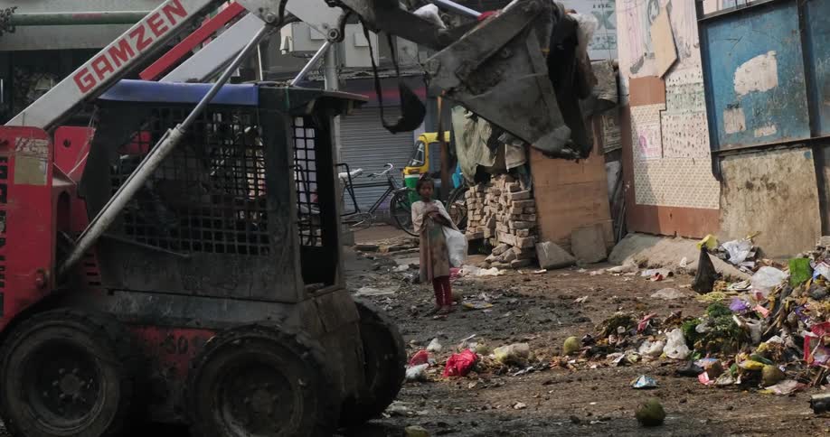 A young girl watches as a bulldozer picks up trash on the street