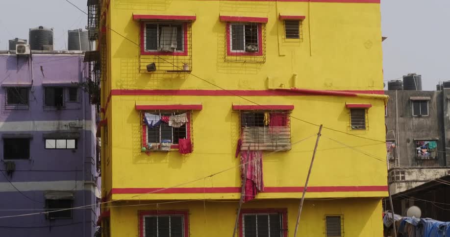 Clothes are hanging outside of the windows of a bright yellow building with red trim with wood and debris in the dirt in front