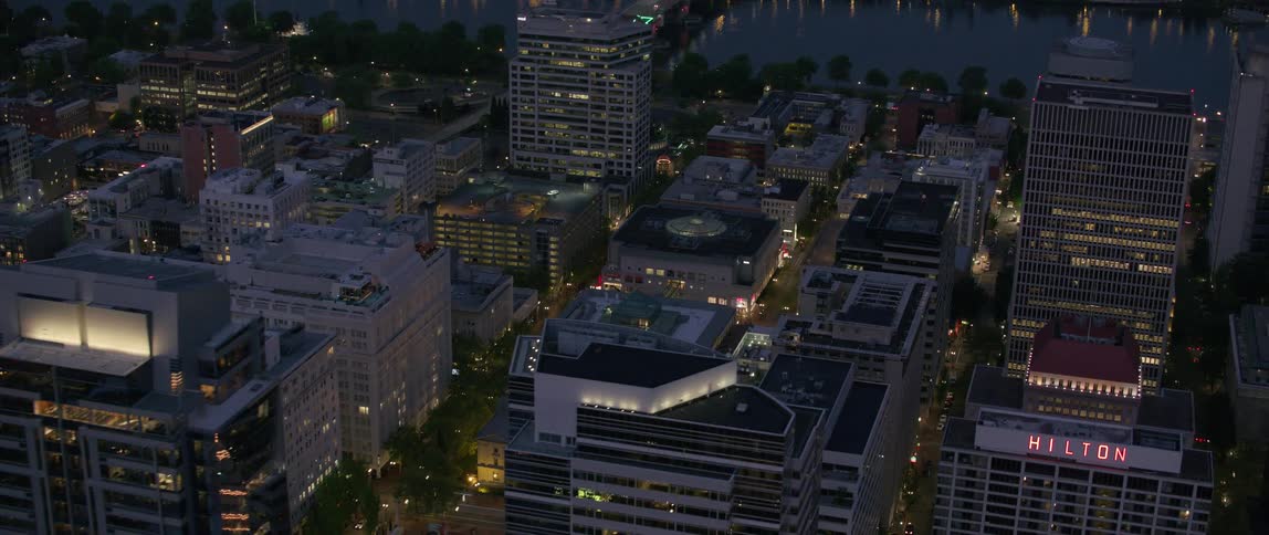 Drone view of city buildings lit up at dusk with a bridge going over water reflecting light