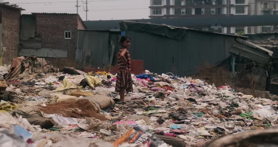 A young girl standing at the top of a large pile of trash in a slum next to a skyscraper
