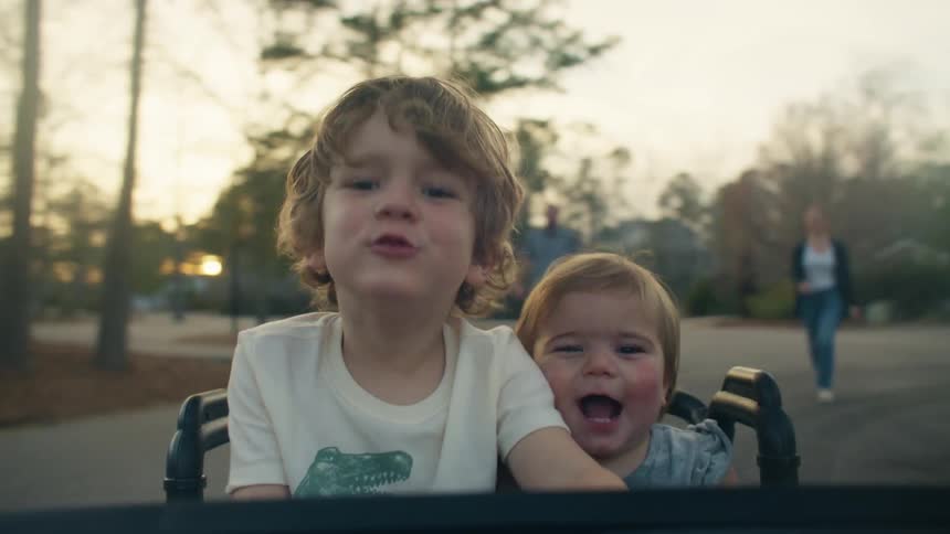 Two young children are laughing and being silly as they ride in a seat on the back of a vehicle on a neighborhood street as two people walk behind them