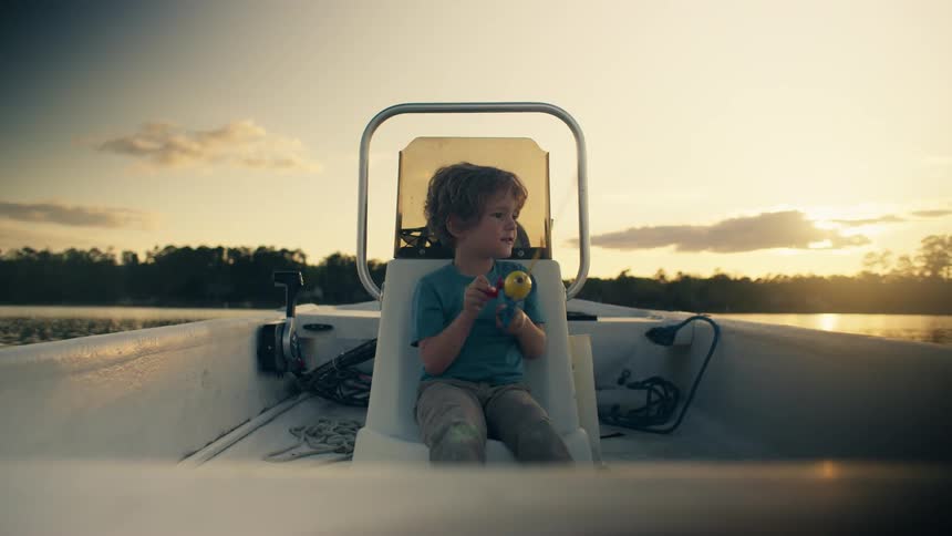 Kid sitting in chair in boat casts fishing line as sun shines from over woods