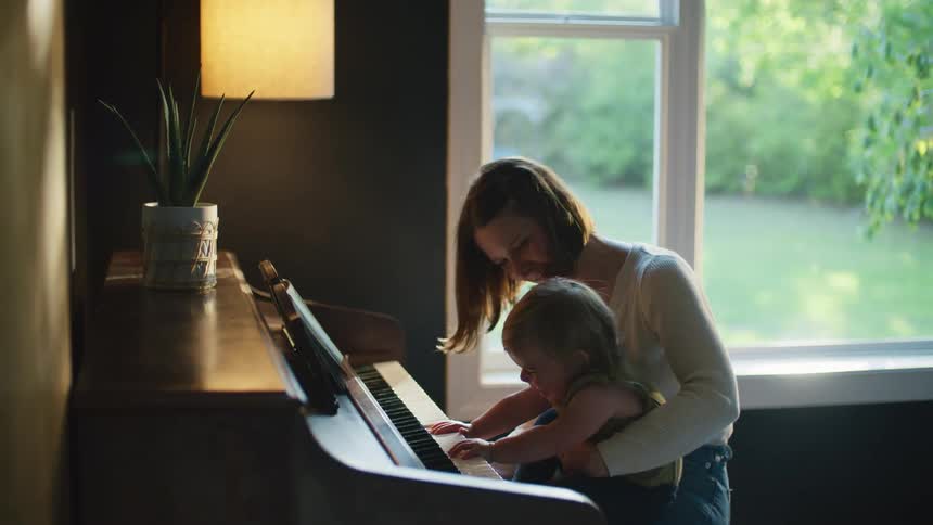 Mother with brown hair white shirt and jeans holds daughter playing piano in room