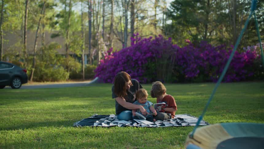 Mother toddler daughter and son sits on outdoor rug on green grass in the yard