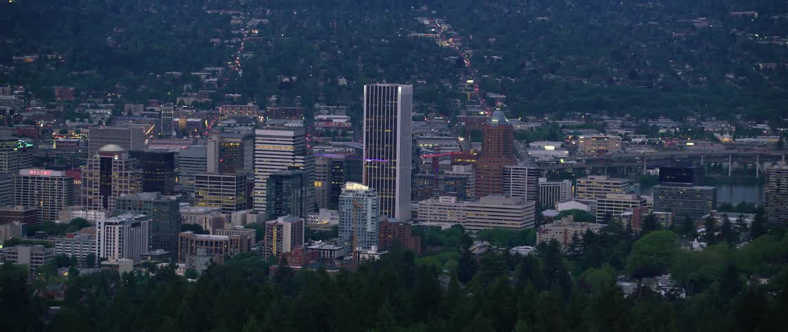An aerial of a city skyline and buildings lit up at dusk with a wide horizon landscape covered in mountains