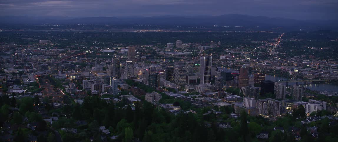 Tall buildings and skyscrapers overlook the city as lights glow and traffic moves on the highway and there are mountains in the distance under a hazy sky