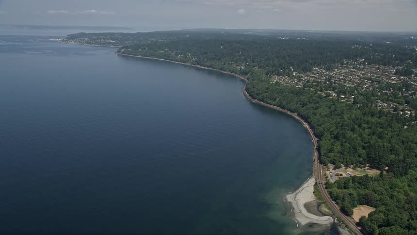 Aerial footage of railroad tracks on the shoreline of Puget Sound with a tilt up to show the Blue Ridge homes by the water in Washington