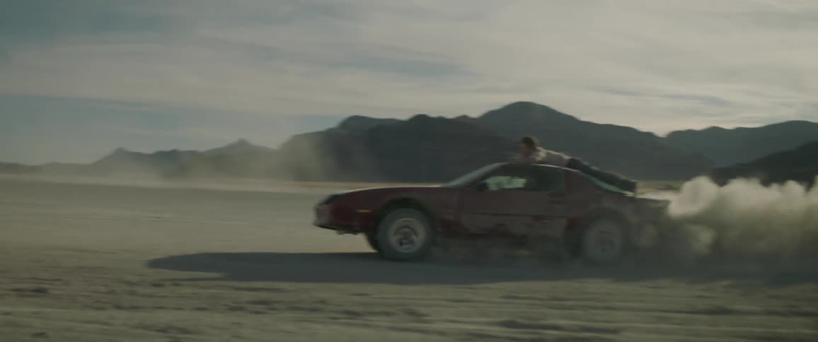 A man is clinging to the top of a car as it speeds in circles in the desert during the day