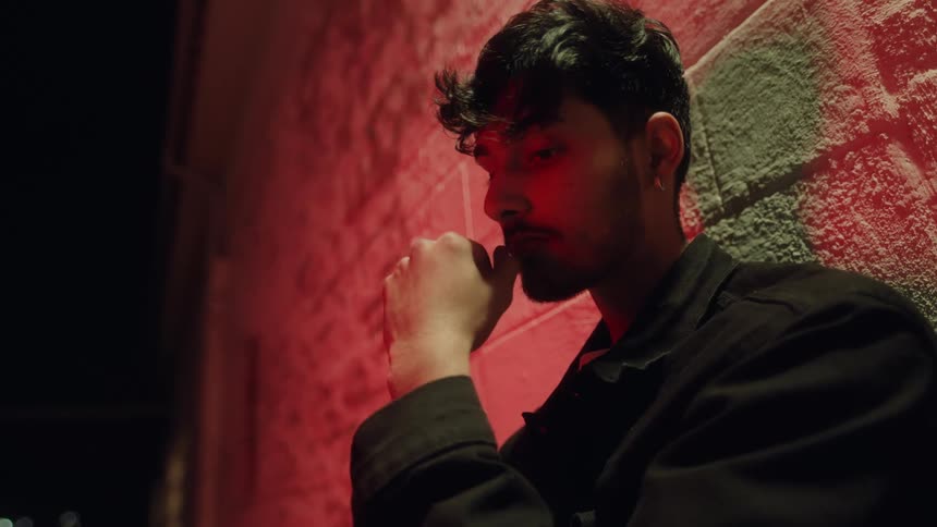 Red light shines on a man standing outside next to a brick wall as he touches his face with his hand