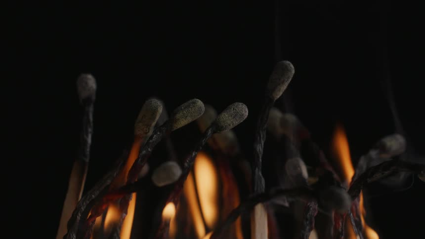 Smoke rises from charred burning cluster of matches in dark area