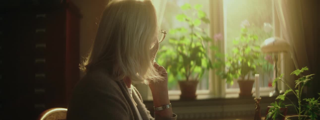 An elderly woman in thought while sitting next to a sunny window at home