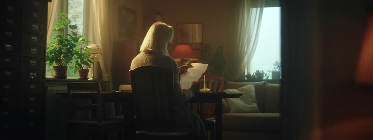 Woman with gray hair jacket and glasses sits at desk reading letter in the living room