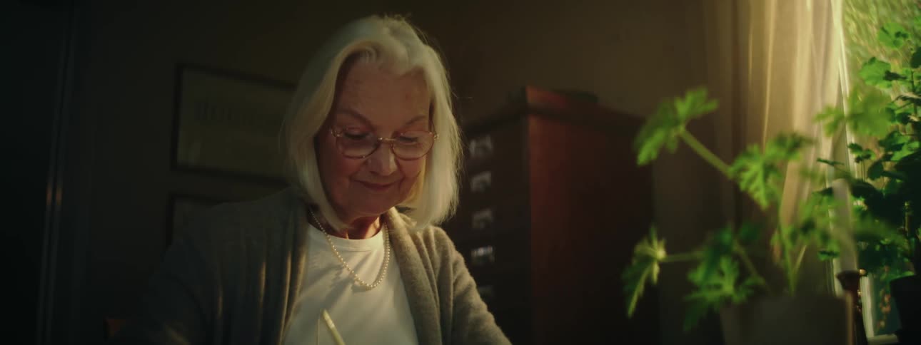 An older woman smiles sentimentally as she writes a letter in an office