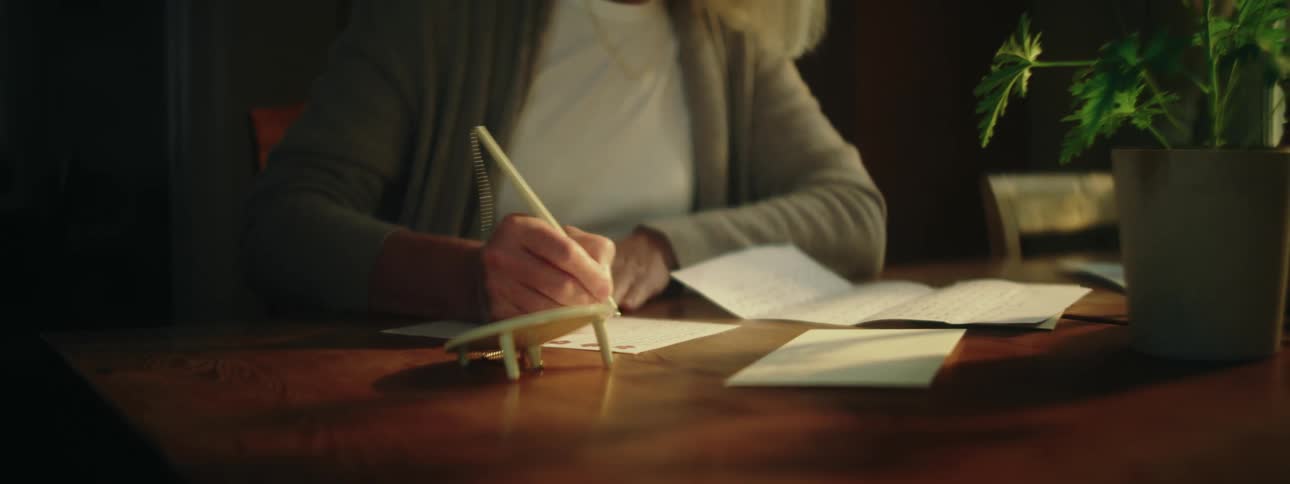 A woman is sitting at a table writing a letter with a smile on her face