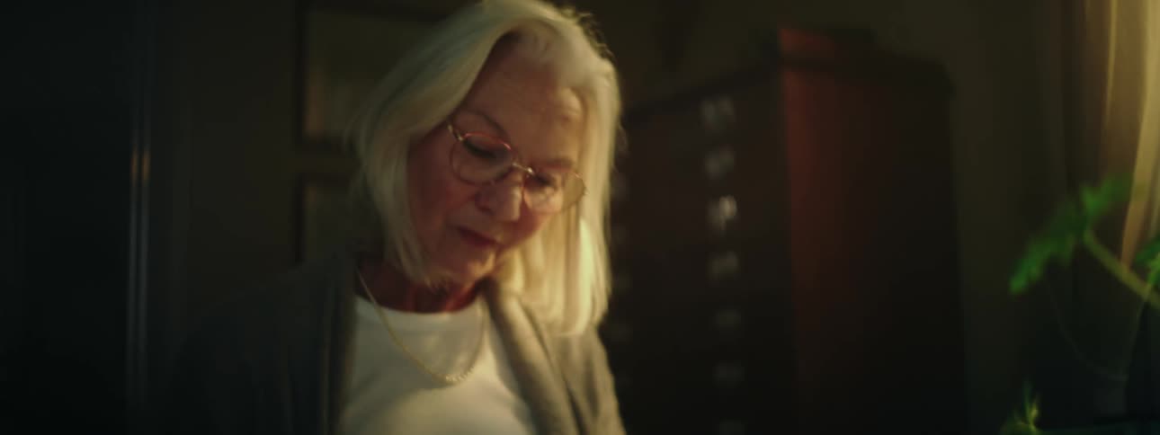 Elderly woman with gray hair glasses and white top sits at desk and writes down a letter