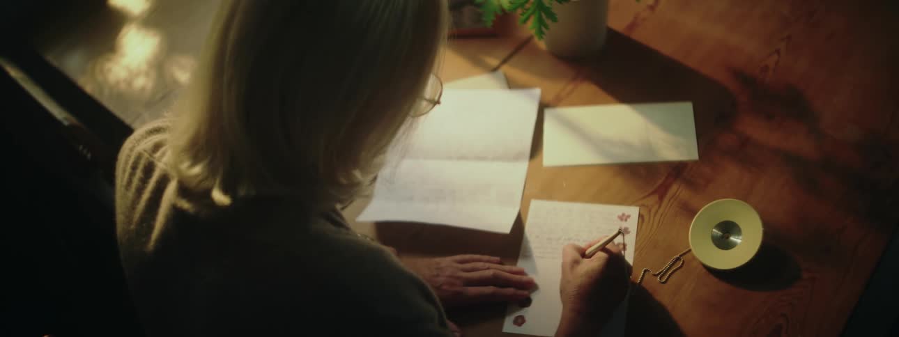 The sun shines brightly upon a desk with plants near a woman as she writes a letter