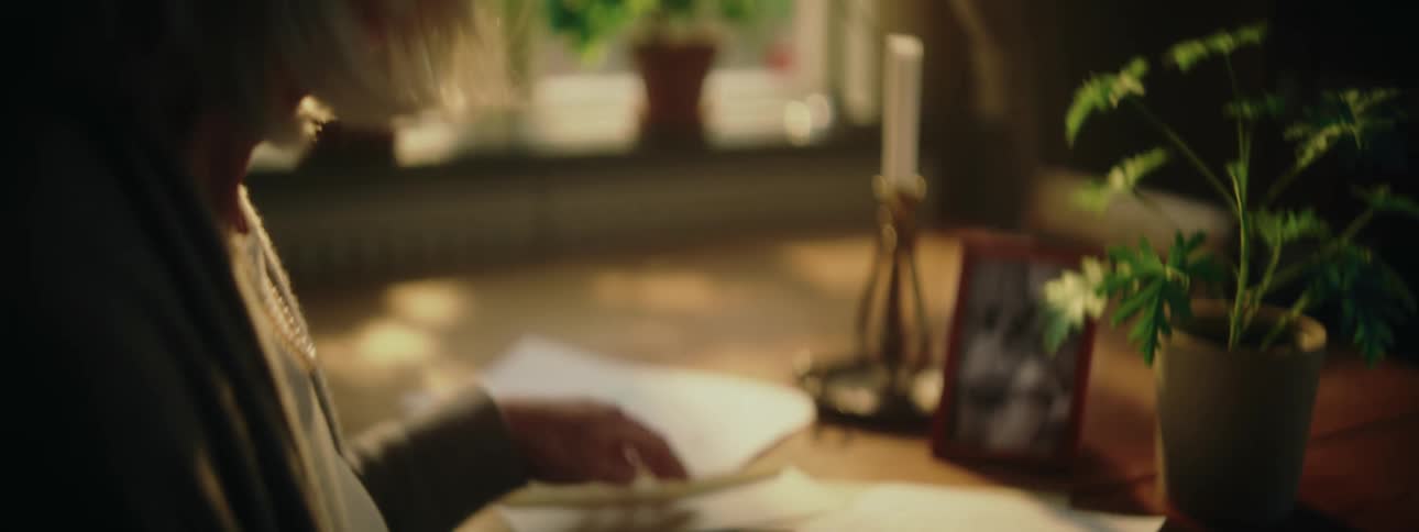A woman is sitting at a desk reading a letter and she puts it in the envelope and picks up a photograph from her desk and holds it with both hands as she looks at it