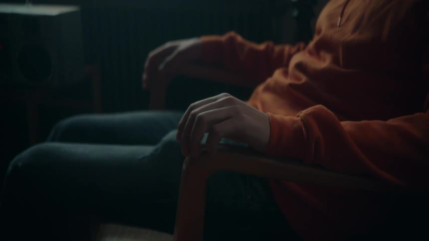 A close up of a person wearing an orange sweatshirt sitting in a chair tapping his fingers on the arm rest