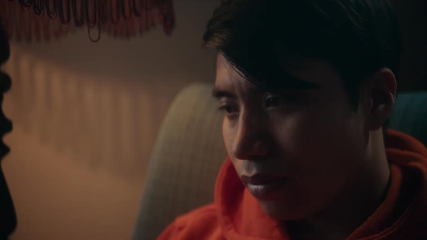 A teenager wearing an orange hoodie is sitting in a chair as light flashes in the room