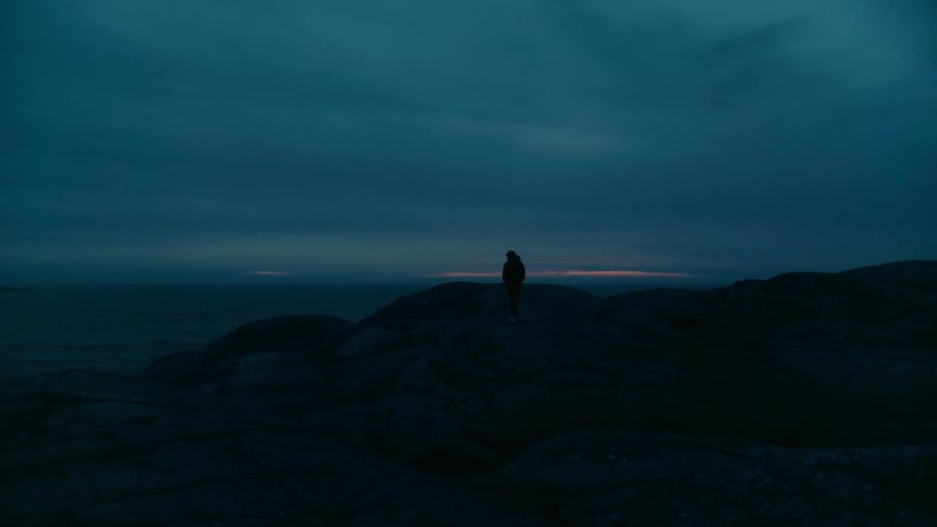A person walks along a rocky surface toward the edge of a cliff overlooking the sea at dusk
