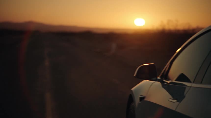 A car drives down a desert road heading toward the sunset