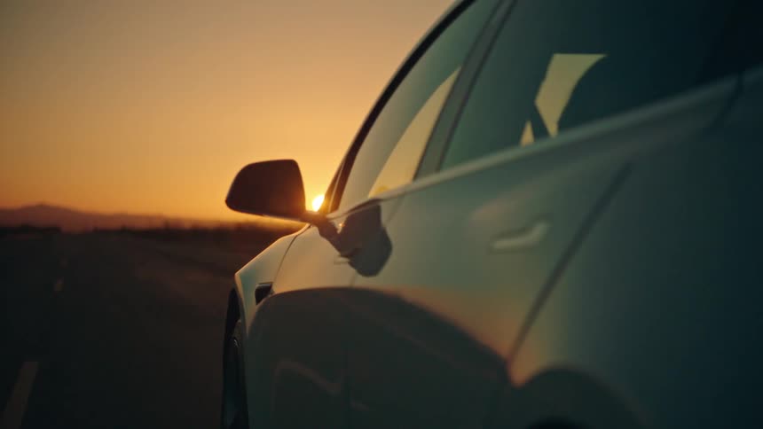 An automobile travels on a road toward a mountain as the orange light from the setting sun fills the sky
